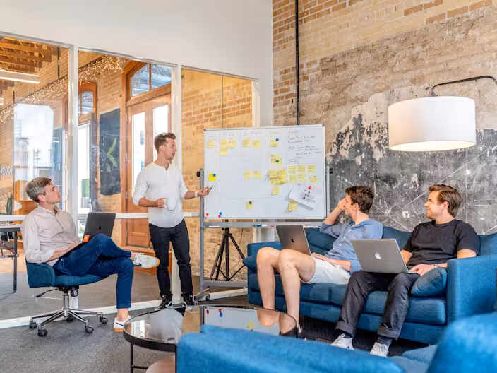 A collaborative team meeting in a modern industrial-style office. One man stands by a whiteboard covered in sticky notes, gesturing toward it, while three colleagues sit on blue sofas and a desk chair with laptops, engaged in the discussion.