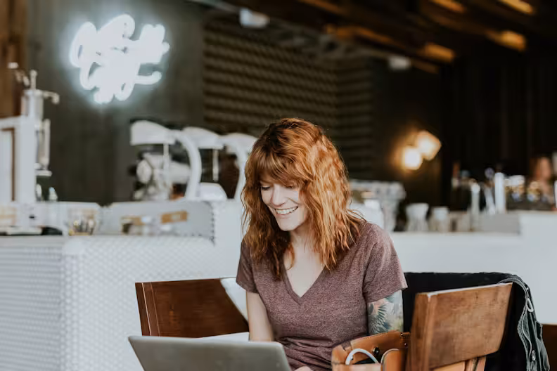 A smiling woman with auburn hair working on a laptop in a bright, modern cafe with a blurred background featuring a neon sign and coffee equipment.