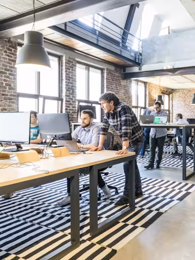 A modern industrial office with brick walls and high ceilings. Two men collaborate at a light wood desk with a monitor, while other colleagues work in the background on a black and white striped rug.