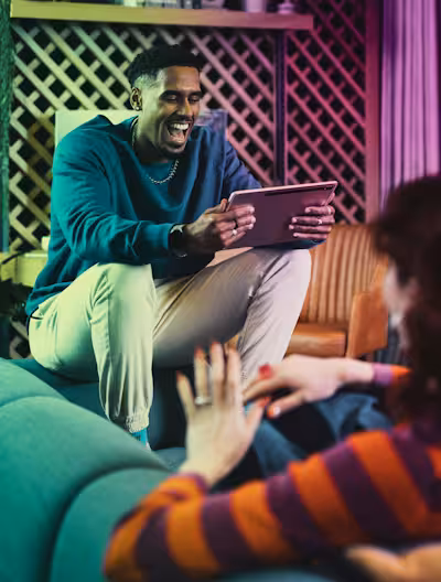 A man sitting on the back of a sofa laughs while holding a tablet, interacting with a woman in a striped sweater. The room is decorated with a wooden lattice wall and warm, colorful mood lighting.