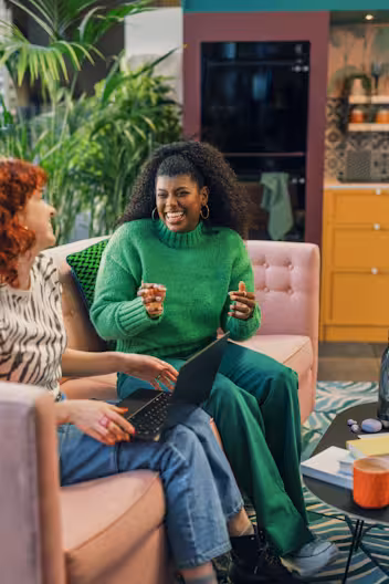 Two women laughing and collaborating on a pink sofa in a colorful modern office. One woman wears a bright green sweater and trousers, while the other sits with a laptop. Large indoor plants and a yellow kitchen area are visible in the background.