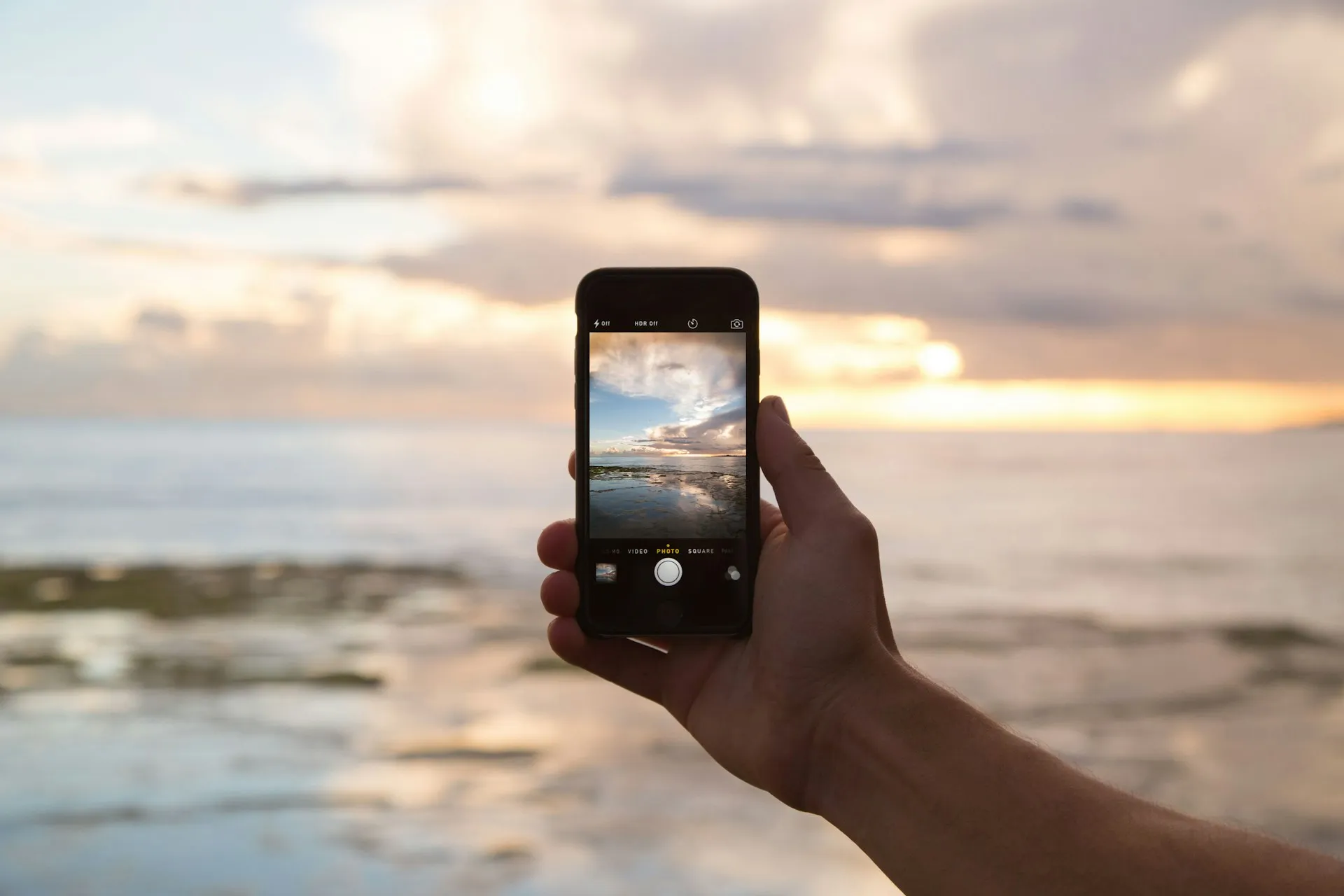 A hand holding up a smartphone to photograph an ocean sunset from a resort balcony, representing the moment a guest is deciding whether to book their stay directly or through a third-party site.