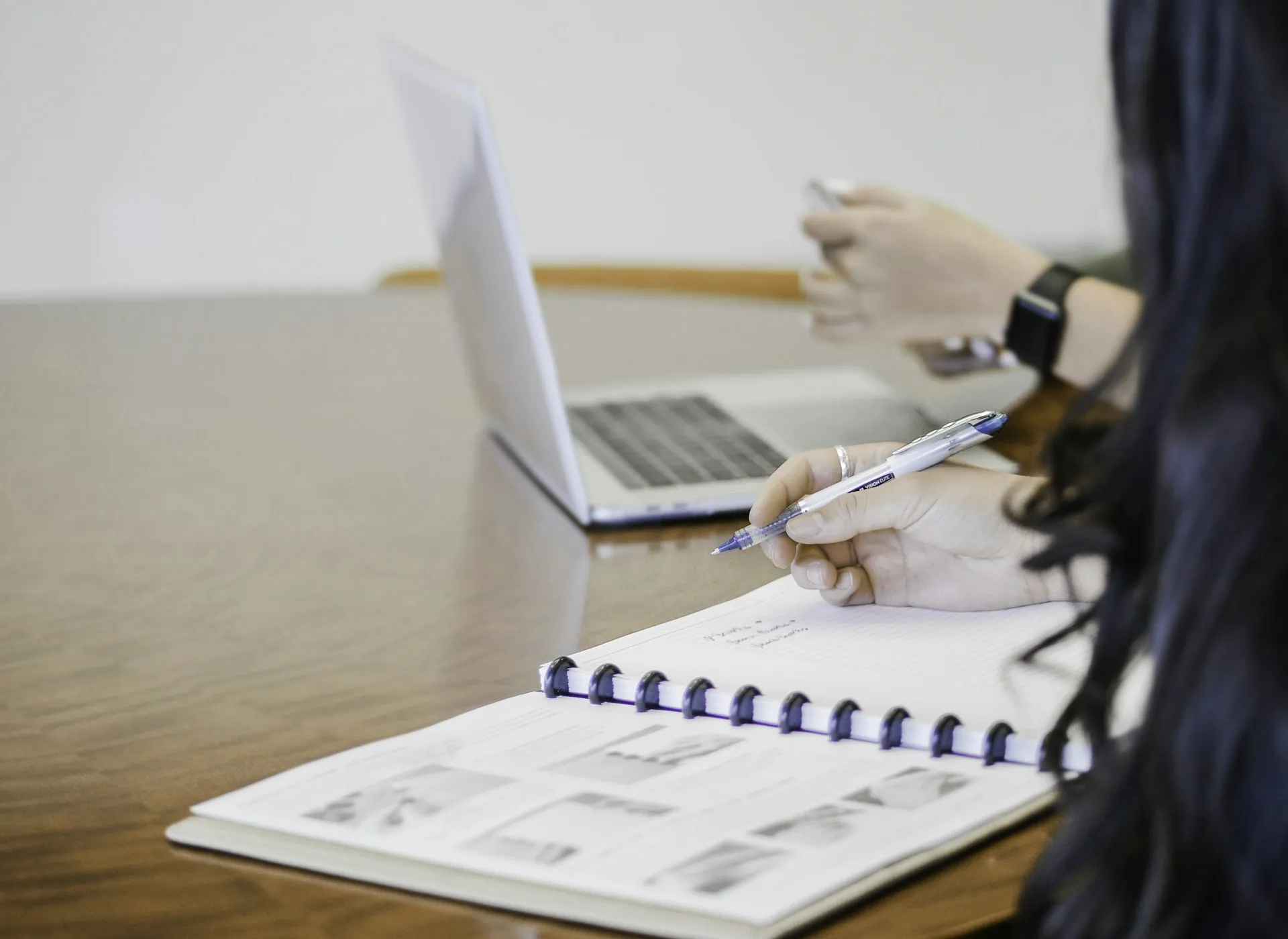 A person writing notes in a notebook next to an open laptop while holding a phone, representing a shopper comparing insurance quotes across multiple tabs and devices.