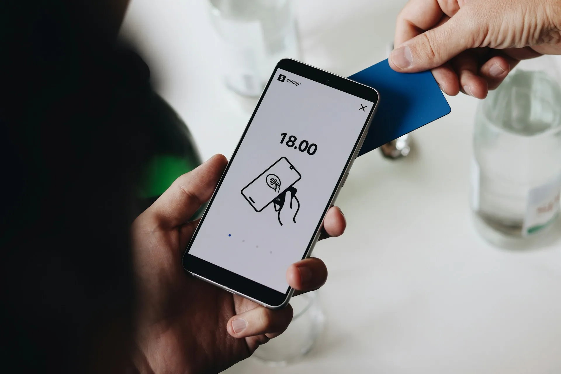 A customer tapping a blue card against a smartphone showing a payment total of 18.00, representing the final price a UK customer sees at checkout.