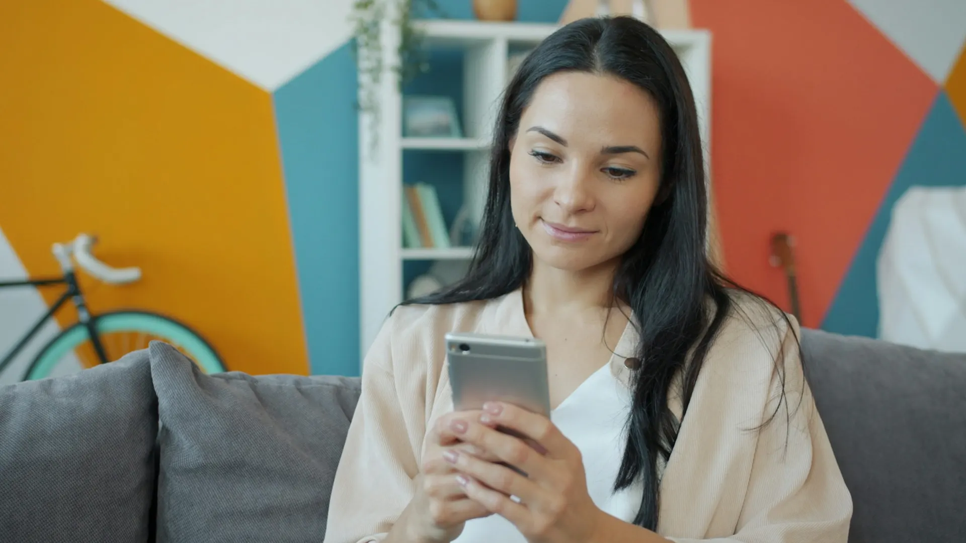 A woman on a sofa in a brightly painted living room looking at her smartphone, representing a customer choosing where to order dinner from on a phone.