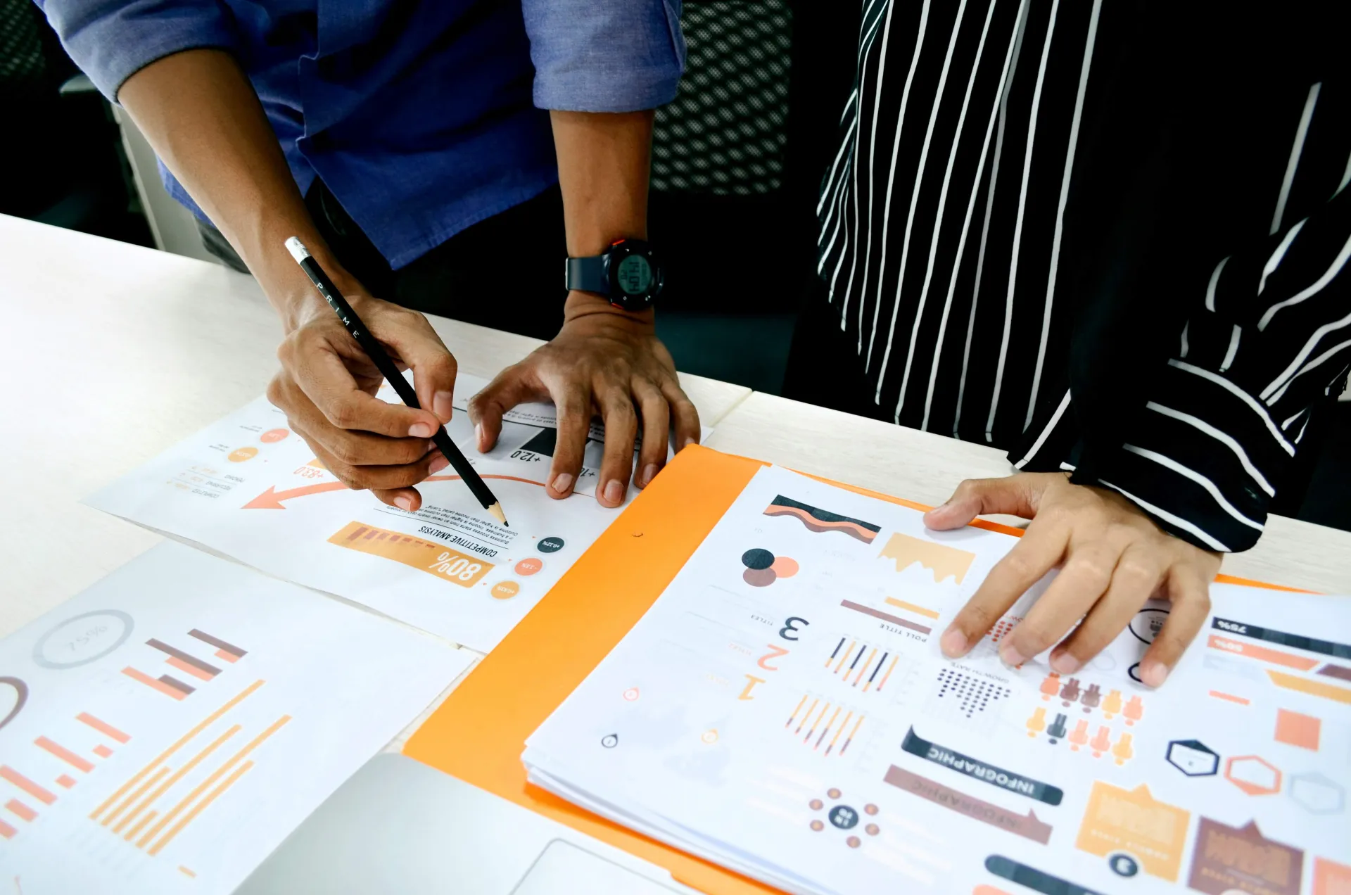 Close-up of two people collaborating on business analytics reports. One person points a pencil at a colorful growth chart on a printed document while another person reviews a stack of infographics in an orange folder.