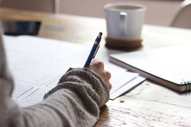 Close-up of a person's hand drawing website wireframes or storyboards with a blue mechanical pencil on a wooden desk, with a coffee mug in the blurred background.