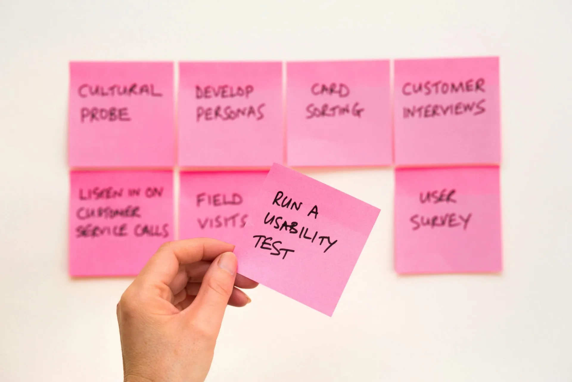 A hand holding a pink sticky note with the text 'Run a usability test' in front of a wall of other UX research methods including card sorting, customer interviews, and user surveys.