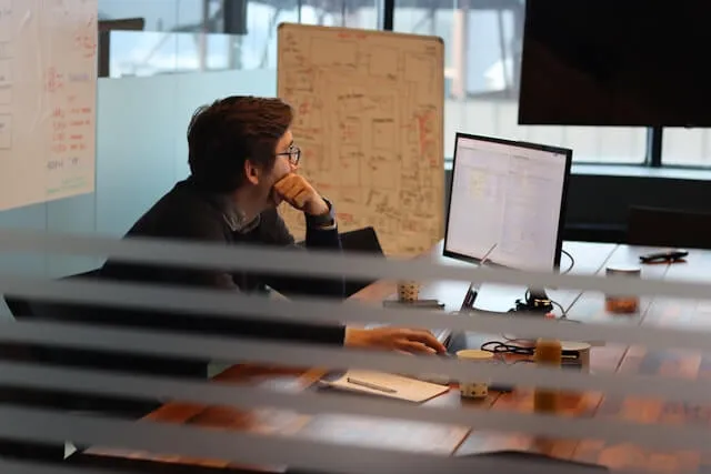 A focused UX researcher analyzing user behavioral data and interface patterns on a computer monitor, with a whiteboard featuring complex user journey maps and wireframe diagrams in the background.