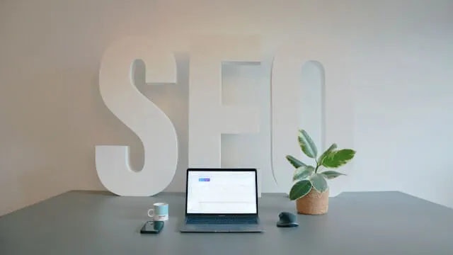 A minimalist, professional workspace featuring an open laptop displaying analytics data, centered in front of large white 3D letters spelling 'SEO' against a clean white wall.